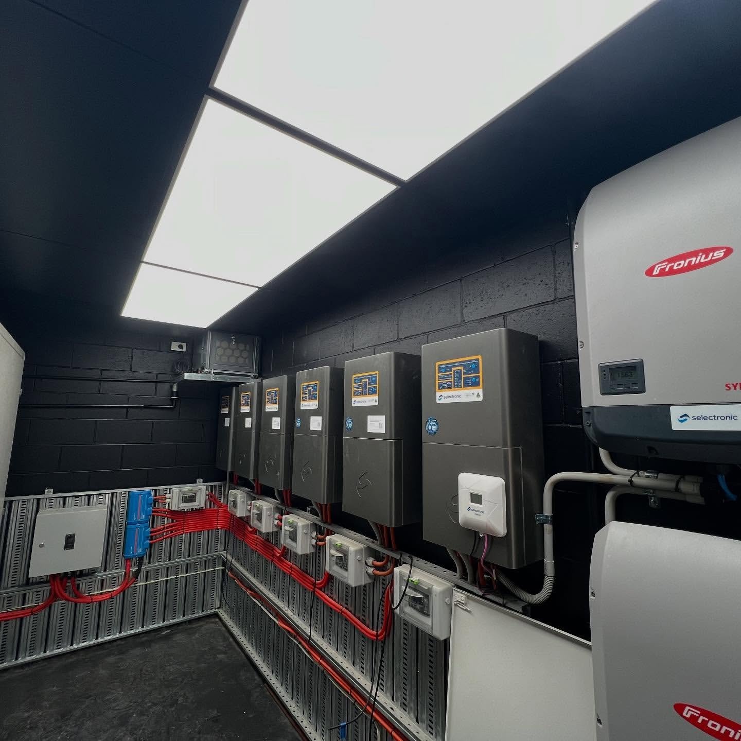 Industrial electrical equipment room with dark panels, mounted inverters, white tanks, and red metal safety barrier