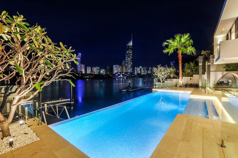 Luxurious waterfront villa with illuminated infinity pool overlooking a city skyline at night