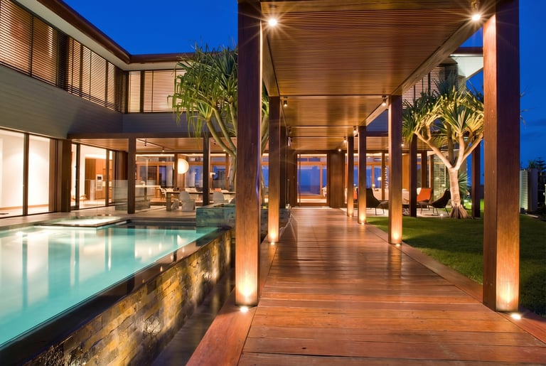 Modern luxury home with illuminated pool, wooden deck, and covered patio at dusk